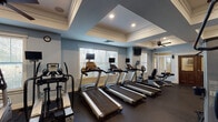 This fitness center offers a comprehensive workout environment designed to accommodate a wide range of exercise needs. The space is divided into distinct functional zones, each thoughtfully arranged to maximize usability and flow. The cardio area features a row of treadmills, each equipped with individual monitor screens, positioned along large windows with white blinds that allow generous natural light to fill the room. A ceiling fan overhead and recessed lighting complement the airy, open atmosphere created by the tray ceilings with crisp white crown molding trim — a refined architectural detail that elevates the overall aesthetic. The walls are finished in a cool, muted blue tone, lending a calm and focused ambiance throughout. The strength training zone includes a Cybex cable machine, adjustable weight benches, and a fully stocked dumbbell rack, offering versatile options for resistance training. A large-screen display is mounted on the wall for visual engagement. A dedicated TRX Training Zone features a full suspension training rig stocked with resistance bands, medicine balls, and accessories, backed by floor-to-ceiling mirrors that enhance the sense of space. Dark rubber flooring runs throughout, providing durability and safety. The facility is accessed through elegant double wood doors with glass panel inserts, reinforcing the polished character of the space.