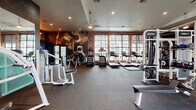 This expansive fitness center is thoughtfully designed to deliver a comprehensive workout experience within a stylish, industrial-inspired setting. The space is anchored by exposed brick walls, dark rubber flooring, and soaring ceilings that create an energetic yet refined atmosphere throughout.

The cardio zone features a row of Life Fitness treadmills and elliptical trainers positioned along large, grid-pane windows that flood the area with natural light and offer views of the surrounding exterior. A dedicated strength training area houses a Synergy 360 functional training system alongside additional cable and resistance machines, providing versatile options for full-body workouts.

The free weights section is well-appointed with a full rack of dumbbells, adjustable benches, and a wall-mounted flat-screen television — ideal for both focused training and entertainment. A bold illuminated "BE FIT" marquee sign adds a motivating focal point above a hydration station along the brick accent wall.

Dramatic large-scale murals depicting athletic imagery adorn the dark feature walls, reinforcing the fitness-focused identity of the space. A teal mesh accent structure adds a pop of color to the weight area. Recessed lighting and pendant fixtures ensure the space is well-lit throughout, while multiple exterior-facing windows and a glass exit door connect the interior to the outdoors.