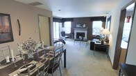 This single-floor residence showcases an open-plan design that seamlessly connects the dining and living areas, creating a spacious and cohesive environment. The dining area is anchored by a dark wood table paired with sleek metal-framed chairs, set atop plush carpeting that extends throughout the space. A decorative floral arrangement adds a refined touch to the table setting.

The living room is defined by a bold, deep brown accent wall that provides a dramatic backdrop for the white-mantled fireplace, which serves as the room's focal point. The fireplace is framed by a clean marble surround, lending an elegant architectural detail to the space. Furnishings include a black leather sofa and contemporary seating, complemented by a glass-topped coffee table.

Multiple large windows line the walls, flooding the interior with abundant natural light and offering views of the surrounding exterior. Dark-toned window treatments provide contrast against the lighter walls and add a layer of privacy. The neutral carpeted floors, combined with the rich color palette and modern furnishings, give the space a sophisticated yet comfortable ambiance throughout.