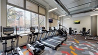 This fitness studio is a thoughtfully designed, single-floor facility that combines functionality with a modern aesthetic. The space is anchored by a row of cardio machines—including treadmills, ellipticals, and a StairMaster—positioned along a wall of large windows fitted with roller shades. These expansive windows flood the studio with natural light while offering views of the surrounding urban streetscape, creating an energizing atmosphere for workouts.

The exposed ductwork and industrial-style ceiling lend the studio a contemporary, loft-like character, complemented by sleek linear pendant lighting. A bold, geometric chevron-patterned carpet in shades of gray, red, orange, and yellow covers much of the floor, adding a vibrant visual element that distinguishes the space.

The strength training area features a cable/pulley machine system, selectorized weight machines, and a dedicated free weights zone complete with a full dumbbell rack and stability balls of varying sizes. Rubberized flooring in this section provides a practical, high-traction surface. Wall-mounted flat-screen televisions are positioned throughout for entertainment during workouts. The studio's open, spacious layout and diverse equipment selection make it a comprehensive facility suited for a wide range of fitness routines.