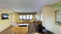 This well-appointed two-story space offers a warm and inviting atmosphere, highlighted by thoughtful design and classic architectural detailing throughout. The main lounge area is generously proportioned, featuring recessed lighting and elegant crown molding that adds a refined touch to the interior. Multiple windows dressed with decorative floral valances and horizontal blinds allow ample natural light to fill the room, creating a bright and pleasant environment.

The lounge is furnished with a curated mix of seating, including plush tan armchairs and rich, dark brown sofas arranged around low coffee tables — providing comfortable areas for conversation or relaxation. A patterned area rug anchors the seating arrangement, adding depth and visual interest to the space. Neutral, warm-toned walls complement the furnishings and contribute to the overall welcoming ambiance.

A set of glass-paned French doors connects to an adjacent room or outdoor area, enhancing the sense of openness and flow between spaces. Architectural columns provide structural definition while maintaining an open, airy feel throughout. The space spans two floors, suggesting additional rooms or amenities beyond what is immediately visible, and is well-suited for communal gathering or hospitality use.