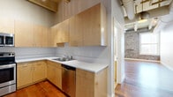 This 2-bedroom, 2-bathroom unit showcases a compelling blend of industrial and modern design elements across a single floor. The open living area immediately draws attention with its soaring ceilings featuring exposed structural beams and large-diameter ductwork, complemented by rich hardwood flooring throughout. Pendant lighting adds an industrial-chic aesthetic to the common spaces.

The kitchen is well-appointed with light maple cabinetry extending to the ceiling, white countertops, and a full suite of stainless steel appliances including a range, over-the-range microwave, dishwasher, and refrigerator. Hardwood flooring continues seamlessly into the kitchen, maintaining a cohesive aesthetic.

The bedrooms feature striking exposed brick accent walls with large windows that allow abundant natural light and exterior views. The primary bedroom offers a comfortable footprint, ceiling fan, and direct hallway access. Both bathrooms are finished with tile flooring, wood-toned vanities, white countertops, and tub-shower combinations with chrome fixtures.

Two hallways connect the rooms efficiently, and a dedicated closet provides additional storage. The overall ambiance balances historic architectural character with clean, modern finishes throughout.