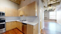 This 2-bedroom, 2-bathroom unit showcases a compelling blend of industrial and modern design elements across a single floor. The open living area immediately draws attention with its soaring ceilings featuring exposed structural beams and large-diameter ductwork, complemented by rich hardwood flooring throughout. Pendant lighting adds an industrial-chic aesthetic to the common spaces.

The kitchen is well-appointed with light maple cabinetry extending to the ceiling, white countertops, and a full suite of stainless steel appliances including a range, over-the-range microwave, dishwasher, and refrigerator. Hardwood flooring continues seamlessly into the kitchen, maintaining a cohesive aesthetic.

The bedrooms feature striking exposed brick accent walls with large windows that allow abundant natural light and exterior views. The primary bedroom offers a comfortable footprint, ceiling fan, and direct hallway access. Both bathrooms are finished with tile flooring, wood-toned vanities, white countertops, and tub-shower combinations with chrome fixtures.

Two hallways connect the rooms efficiently, and a dedicated closet provides additional storage. The overall ambiance balances historic architectural character with clean, modern finishes throughout.