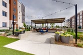Located at 3920 N Salida St in Aurora, Colorado, this well-appointed outdoor amenity space offers a thoughtfully designed gathering area for residents and guests. The space is divided into two distinct zones that work seamlessly together. A generous artificial turf lawn provides a vibrant, low-maintenance green area ideal for recreation and relaxation, bordered by landscaped planting beds featuring ornamental grasses, flowering plants, and decorative gravel. An elegant wrought iron perimeter fence defines the space while maintaining open sightlines to the surrounding neighborhood. The hardscaped patio area is anchored by a substantial steel pergola with a corrugated roof panel, offering partial shade and shelter. Beneath it, built-in brick grilling stations are positioned on either side, making the area well-suited for outdoor cooking and entertaining. A round dining table with vibrant blue chairs sits centrally under the pergola, creating an inviting al fresco dining setting. Large modern planters filled with colorful blooms accent the patio edges, while string lights crisscross overhead across the entire courtyard, lending a warm, festive ambiance during evening hours. The overall design balances functional amenities with aesthetic appeal, creating a versatile outdoor destination.