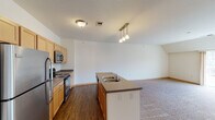 This single-floor unit is designed with an open-concept layout that connects the kitchen and living areas, creating a spacious and functional flow throughout. The kitchen is outfitted with light maple cabinetry, dark laminate countertops, and stainless steel appliances including a refrigerator, electric range, and over-the-range microwave. A central island with a double basin sink and pendant lighting above serves as both a practical workspace and a natural dividing point between the kitchen and the expansive carpeted living area. Hard wood-style flooring runs through the kitchen and entry zones, transitioning to plush carpet in the living and bedroom areas. The living space is generously sized with vaulted ceiling lines that add architectural interest and a sense of openness. Bedroom areas feature soft carpet, closet storage with hanging rods, and windows that allow natural light to filter in. The bathroom includes a vanity with matching cabinetry, a full soaking tub with a curved shower rod, vinyl plank flooring, and grab bars along the walls. Clean white walls, warm wood trim, and consistent finishes throughout give the unit a cohesive, move-in ready appearance.