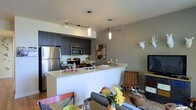 This single-floor space showcases a thoughtfully designed open-concept layout that integrates the kitchen, dining, and living areas into one cohesive environment. The kitchen is outfitted with stainless steel appliances, including a refrigerator, microwave, and range, complemented by dark wood cabinetry that provides a striking contrast against the light-toned countertops. A peninsula-style breakfast bar extends from the kitchen, serving as both a functional workspace and a natural divider between the kitchen and living areas, while also offering casual seating possibilities. Pendant lighting — including a cylindrical white fixture and a sleek tubular pendant — hangs above the counter, adding warmth and visual interest. A track lighting system further illuminates the kitchen zone. The living area is comfortably appointed with a gray sofa, decorative accents, and a wall-mounted flat-screen television. A white decorative animal head mounted on the taupe-toned accent wall adds a touch of eclectic personality to the space. The overall ambiance is modern and urban, with a neutral color palette punctuated by warm wood tones and carefully chosen décor elements that create an inviting, stylish atmosphere.