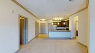 This single-floor residential unit offers a thoughtfully designed open-concept layout that seamlessly connects the living area and kitchen. The kitchen is a standout feature, equipped with dark wood cabinetry, granite countertops, and stainless steel appliances including a refrigerator, microwave, and range. A peninsula island with an undermount sink provides additional prep space and serves as a natural divider between the kitchen and the living area. Elegant pendant lighting hangs above the island, adding a warm, modern touch to the space. Crown molding trims the ceilings throughout, lending a refined architectural detail to the interiors. The living area features plush carpeting, while the kitchen and entryway transition to hard surface flooring, creating a practical and visually appealing contrast. Multiple doors visible from the main living space suggest well-defined private rooms, offering both functionality and privacy. The neutral color palette of soft whites and warm grays throughout creates a clean, welcoming ambiance. The layout maximizes usability and flow, making this a comfortable and well-appointed single-level living space.