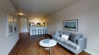 This well-appointed single-floor space offers a thoughtfully designed open-concept layout that effortlessly blends living and kitchen areas into one cohesive environment. The living area is anchored by a plush gray sofa complemented by a sleek round white coffee table, creating a comfortable and inviting atmosphere. Warm hardwood flooring runs throughout the space, adding a rich, natural tone that contrasts beautifully with the neutral gray walls. A framed landscape artwork adds a touch of color and character to the living area. The kitchen is outfitted with white cabinetry, stainless steel appliances — including a refrigerator and microwave — and is illuminated by modern track lighting that highlights the clean, functional workspace. A peninsula with bar-height seating accommodates three stools, making it ideal for casual dining or entertaining. The overall ambiance is bright, modern, and welcoming, with a clean aesthetic that balances practicality and style. The open flow between spaces makes the layout feel expansive and versatile, well-suited for both everyday living and hosting guests.