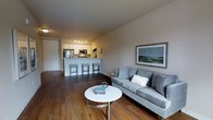 This well-appointed single-floor space offers a thoughtfully designed open-concept layout that effortlessly blends living and kitchen areas into one cohesive environment. The living area is anchored by a plush gray sofa complemented by a sleek round white coffee table, creating a comfortable and inviting atmosphere. Warm hardwood flooring runs throughout the space, adding a rich, natural tone that contrasts beautifully with the neutral gray walls. A framed landscape artwork adds a touch of color and character to the living area. The kitchen is outfitted with white cabinetry, stainless steel appliances — including a refrigerator and microwave — and is illuminated by modern track lighting that highlights the clean, functional workspace. A peninsula with bar-height seating accommodates three stools, making it ideal for casual dining or entertaining. The overall ambiance is bright, modern, and welcoming, with a clean aesthetic that balances practicality and style. The open flow between spaces makes the layout feel expansive and versatile, well-suited for both everyday living and hosting guests.