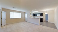 This single-floor space showcases an open-concept design that seamlessly connects the living area to the kitchen. The living area is finished with plush, neutral-toned carpeting, creating a comfortable and cohesive feel throughout. A large window along one wall allows natural light to fill the space, brightening the warm beige walls and contributing to an airy atmosphere. A white-paneled exterior door nearby adds a secondary source of natural light.

The kitchen is defined by striking dark cabinetry that contrasts sharply against the light walls and white appliances, including a built-in microwave and a range. A peninsula with a light-toned countertop serves as a natural divider between the kitchen and living area, offering additional workspace and potential seating. Recessed lighting and a skylight above the kitchen ensure the workspace remains well-lit. The kitchen floor transitions to a hard surface, providing a practical distinction from the carpeted living space. A hallway visible beyond the kitchen suggests additional rooms within the unit, further enhancing the functional layout of this well-designed space.
