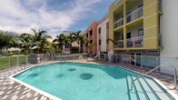This outdoor amenity space showcases a generously sized, irregularly shaped swimming pool with crystal-clear turquoise water, complete with built-in bench seating and a secure perimeter railing. The pool deck is finished with attractive paver tiles, providing a clean and functional surround ideal for lounging and recreation. Lush, mature tropical palm trees frame the area, enhancing the resort-like atmosphere and offering natural shade throughout the day.

The backdrop is defined by a vibrant multi-story residential building featuring a striking color palette of coral, white, and yellow-green tones. Each floor is equipped with private balconies enclosed by metal railings, adding architectural dimension and a sense of vertical openness to the overall design. A safety ring station is conveniently positioned along the pool fence, reflecting attention to resident safety.

The overall ambiance is bright, airy, and tropical, with an open sky allowing ample natural light to fill the space throughout the day. The combination of colorful architecture, swaying palms, and inviting pool waters creates a lively, vacation-inspired setting well-suited to warm-weather living. This communal outdoor space serves as a central gathering point, offering residents a refreshing retreat from daily life.