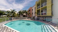This outdoor amenity space showcases a generously sized, irregularly shaped swimming pool with crystal-clear turquoise water, complete with built-in bench seating and a secure perimeter railing. The pool deck is finished with attractive paver tiles, providing a clean and functional surround ideal for lounging and recreation. Lush, mature tropical palm trees frame the area, enhancing the resort-like atmosphere and offering natural shade throughout the day.

The backdrop is defined by a vibrant multi-story residential building featuring a striking color palette of coral, white, and yellow-green tones. Each floor is equipped with private balconies enclosed by metal railings, adding architectural dimension and a sense of vertical openness to the overall design. A safety ring station is conveniently positioned along the pool fence, reflecting attention to resident safety.

The overall ambiance is bright, airy, and tropical, with an open sky allowing ample natural light to fill the space throughout the day. The combination of colorful architecture, swaying palms, and inviting pool waters creates a lively, vacation-inspired setting well-suited to warm-weather living. This communal outdoor space serves as a central gathering point, offering residents a refreshing retreat from daily life.