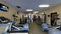 This fitness center offers a comprehensive workout environment designed to accommodate a variety of exercise routines. The space is laid out in a long, corridor-style floor plan, allowing for an organized arrangement of equipment along both sides. Precor elliptical trainers and cardio machines are prominently featured in the foreground, complemented by a full selection of selectorized strength machines arranged toward the center and rear of the room. Multiple wall-mounted flat-screen televisions are positioned throughout the space, providing entertainment while exercising. The flooring consists of durable, textured rubber matting suited for high-traffic fitness use. Overhead fluorescent lighting runs the length of the ceiling, ensuring the space is well-lit throughout. A textured ceiling and neutral-toned walls contribute to a clean, functional aesthetic. A water cooler is conveniently accessible near the entrance. Large mirrors are incorporated along the walls toward the back, enhancing the sense of depth and allowing users to monitor their form. The overall layout is practical and spacious, providing ample room to move between equipment comfortably.