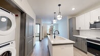 This thoughtfully designed one-bedroom, one-bathroom residence in Jacksonville, Florida offers a contemporary living experience with a clean, modern aesthetic throughout. The open-concept kitchen and living area serves as the heart of the home, anchored by a striking central island with a white quartz countertop, an undermount sink, and a matte black faucet. Stylish pendant lights hang above the island, complementing the two-toned cabinetry — white upper cabinets paired with gray lower cabinets — and a geometric tile backsplash. Stainless steel appliances complete the well-appointed kitchen. Light wood-look flooring flows seamlessly through the main living areas, creating a bright and cohesive feel. The bedroom features plush carpeting, a ceiling fan, and dual windows that invite natural light and outdoor views. The full bathroom is finished with a gray vanity, white quartz countertop, a framed mirror, and a glass-door shower. It connects directly to a spacious walk-in closet outfitted with built-in shelving and hanging rods for ample storage. A dedicated laundry area adds everyday convenience, rounding out this efficiently laid-out and modern residence.