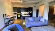 This single-floor loft showcases a compelling industrial design that blends raw architectural elements with functional living spaces. The open-concept layout connects the kitchen and living area in one cohesive environment, creating a spacious and airy feel throughout.

The kitchen is outfitted with sleek, dark cabinetry that runs floor to ceiling, providing generous storage space. A full suite of appliances, including a refrigerator, microwave, and range, are seamlessly integrated into the cabinetry. A central kitchen island with a light-colored countertop serves as a practical workspace and casual dining area, complemented by bar-height seating.

The living area flows naturally from the kitchen and features polished concrete floors that anchor the industrial aesthetic. The space comfortably accommodates a large sectional-style sofa arrangement, making it well-suited for both relaxation and entertaining. A washer and dryer unit is conveniently housed within the main living space.

Overhead, exposed structural beams and a track lighting system add to the loft's industrial character while providing flexible illumination throughout. The open ceiling height contributes to the overall sense of volume and openness. The combination of dark cabinetry, concrete floors, and exposed architectural details gives the space a modern, urban ambiance.
