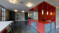 This single-floor commercial space makes an immediate visual impact through its bold, contemporary design. The focal point of the space is a striking red reception or service counter, featuring a solid red laminate surface and frosted acrylic lower panels that diffuse light subtly. A matching red overhead soffit frames the counter area, reinforcing the space's strong color identity and helping define distinct functional zones within the open floor plan.

Overhead, a series of cylindrical pendant lights hang at varying lengths above the counter, complementing flush-mount dome fixtures that illuminate the broader open area. The combination creates layered, well-distributed lighting throughout the space. Underfoot, polished concrete flooring extends across the entirety of the space, lending an industrial yet refined character.

The rear of the space transitions into a secondary area furnished with chairs and tables, suggesting a waiting or lounge zone. Dark accent walls contrast effectively against white walls, adding depth and visual interest. Glass double doors at the entry allow natural light to filter in while offering a transparent connection to the exterior. The overall ambiance is modern, energetic, and professionally designed.
