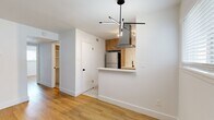 This thoughtfully updated 2-bedroom, 1-bathroom unit offers a clean, modern aesthetic across its single-floor layout. The open-concept living and kitchen area serves as the heart of the home, where warm wood cabinetry, white quartz countertops, and a striking stainless steel island range hood create a polished, contemporary look. The kitchen is well-appointed with stainless steel appliances — including a full-size refrigerator, gas range, and dishwasher — complemented by a tiled backsplash and a peninsula that separates the kitchen from the living area. Light hardwood flooring flows through the living spaces, contrasting with the clean white walls and adding warmth to the overall aesthetic. Natural light enters through horizontal-blind windows throughout the unit. Both bedrooms are well-proportioned, with the primary bedroom offering generous space, a ceiling fan, and a window with exterior views. The second bedroom mirrors a similar clean, bright character. The bathroom features large-format tile flooring, a wood-toned vanity with a quartz countertop, and matte black hardware accents. A hallway with open shelving provides additional storage, connecting the rooms efficiently.