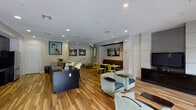 Located on Burgoyne Road in Houston, Texas, this single-floor community amenity space offers a well-appointed, contemporary environment designed for both relaxation and casual gathering. The open-concept layout is unified by warm-toned hardwood flooring that runs throughout, complementing the neutral gray walls and crisp white ceilings fitted with recessed lighting.

The main lounge area is furnished with comfortable upholstered seating in muted green and teal tones, arranged around low accent tables and anchored by a flat-screen television mounted on a textured feature wall. Large windows and glass double doors flood the space with natural light and provide direct views of an outdoor pool courtyard, creating a seamless connection between indoor and outdoor areas.

A dining nook with bright green chairs and compact tables is positioned to maximize the pool views through a large picture window. The kitchenette is sleekly outfitted with dark cabinetry, mosaic tile backsplash, a stainless steel refrigerator, and dual-basin sink. A front counter area with dark countertops serves as a reception or service point. Contemporary artwork accents the walls throughout, contributing to the polished, modern ambiance of the overall space.