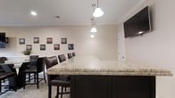 This single-floor space is thoughtfully designed to offer a welcoming and functional environment. The centerpiece of the space is a generously sized bar or counter area finished with a striking light granite surface, set atop dark espresso-toned cabinetry that provides a sharp visual contrast. A row of dark leather bar stools lines the counter, offering comfortable seating for multiple guests. Overhead, a trio of elegant pendant lights hang in a cascading arrangement, casting warm, focused illumination across the counter surface. The neutral, light-toned walls are accented by crown molding that runs along the ceiling perimeter, lending a polished, refined character to the interior. A curated selection of framed artwork is displayed along one wall, adding personality and visual interest to the space. The flooring features a subtle patterned carpet that complements the overall color palette. A wall-mounted screen is positioned in the corner, suggesting the space is suited for both social gatherings and presentations. The overall ambiance is clean, modern, and inviting, making it well-suited for entertaining, casual meetings, or relaxed gatherings.
