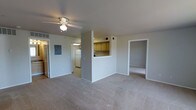 This single-floor residence features an open-concept layout that connects the living area to the kitchen and additional rooms. The living space is finished with neutral wall-to-wall carpeting and smooth white ceilings, creating a clean and understated aesthetic throughout.

A ceiling fan with an integrated light fixture is centrally positioned in the main living area, providing both air circulation and ambient lighting. The space flows naturally toward a pass-through kitchen, which is partially separated by a half-wall counter. The kitchen features warm-toned wood cabinetry, a white refrigerator, and recessed under-cabinet lighting, providing a compact but practical cooking space. An electrical panel is visibly mounted on the wall near the kitchen entry, a notable utilitarian feature.

To the right of the living area, an open doorway leads to an additional room finished with the same neutral carpeting, offering flexible use as a bedroom or flex space. The overall design prioritizes functionality with a straightforward, practical floor plan. The neutral color palette throughout lends itself to easy personalization and a move-in-ready feel.
