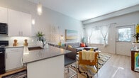 This thoughtfully designed single-floor residence offers a seamless blend of modern aesthetics and functional living. The open-concept layout connects the kitchen and living area, creating an inviting, airy atmosphere filled with natural light from large windows dressed in sheer white curtains. The living area features light wood flooring, a comfortable sectional sofa, and a vibrant chevron-patterned area rug that adds a pop of color and personality to the space. A large statement artwork anchors the living room wall, complementing the overall contemporary design. The kitchen is outfitted with sleek white cabinetry extending to the ceiling, black stainless steel appliances including a gas range, microwave, and refrigerator, and a light tile backsplash. A peninsula with a dark quartz countertop provides additional prep space and serves as a casual dining area with bar seating. Pendant lighting adds a refined touch overhead. The bedroom is a calm, well-proportioned retreat, benefiting from natural light through a window with sheer drapes and featuring ample built-in storage above the bed. The bathroom presents a clean, modern finish with a dark wood-framed vanity, light stone countertop, and wood-look flooring, completing the cohesive design language found throughout the space.