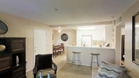 This single-floor residence offers a well-designed open-concept layout that brings together the living, dining, and kitchen areas in one cohesive space. The living area is carpeted and comfortably sized, providing a relaxed setting anchored by warm neutral tones on the walls and ceiling. A ceiling fan adds both function and a casual aesthetic to the space.

The kitchen is positioned prominently at the heart of the home, featuring a peninsula countertop with bar-height seating — ideal for casual dining or entertaining. White cabinetry lines the kitchen walls, complemented by a skylight that draws in natural light and brightens the space. A full-size refrigerator and ample counter space make this kitchen as practical as it is inviting.

Just beyond the kitchen, a dedicated dining area provides a more formal setting for meals, furnished with a wooden dining table and chairs. Decorative wall art adds a personal touch to the transition between spaces.

Throughout the home, the consistent use of neutral carpet, soft wall tones, and white trim creates a clean, unified ambiance. The open sightlines between areas enhance the sense of space and make the floor plan feel both airy and connected — well-suited for everyday living.