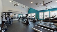 This fitness center is designed to support a comprehensive workout experience within a thoughtfully arranged, single-floor layout. The space is immediately distinguished by its soaring vaulted ceilings, which create an open, airy atmosphere enhanced by multiple ceiling fans that promote air circulation throughout. Bold teal accent walls contrast with crisp white surfaces, giving the gym a modern, energizing aesthetic.

The main workout area is outfitted with a full row of treadmills and elliptical machines, ideal for cardiovascular training, along with a multi-station cable and weight machine for strength exercises. A dedicated free weights section features a well-stocked dumbbell rack, weight benches, and a squat/barbell rack, catering to a wide range of strength training needs. Floor-to-ceiling mirrors line the walls, visually expanding the space while allowing users to monitor their form.

Durable rubber flooring covers the entire workout area, providing safety and support underfoot. Natural light streams in through multiple windows, offering views of the surrounding exterior. A wall-mounted flat-screen television is positioned for easy viewing during cardio sessions. Additional amenities include a water fountain station and a conveniently located restroom with a distinctively designed glass-panel wood door. Recessed lighting and ceiling fans ensure the environment remains comfortable throughout the day.