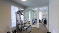 Located at 3202 W Nine Mile Rd. in Pensacola, Florida, this single-floor facility is designed to support both physical fitness and relaxation. The workout area is well-appointed, featuring a Hoist multi-station cable machine as its centerpiece, ideal for a comprehensive full-body training regimen. The room is finished with durable carpeted flooring and painted in a neutral gray palette, contributing to a clean, focused environment. Multiple windows fitted with white horizontal blinds allow natural light to fill the space while maintaining privacy. Adjoining the fitness area through large glass-paned French doors is a dedicated relaxation room, which adds a wellness-oriented dimension to the facility. This secondary space features a colorful area rug, comfortable seating, and décor accents including a prominent "RELAX" sign, creating a calm and inviting atmosphere. A bar-height stool and additional accent pieces suggest the room may also function as a lounge or recovery area. The open sightlines between the two rooms, enabled by the floor-to-ceiling glass partition, create a sense of connectivity and spaciousness throughout the facility. Together, the spaces offer a well-rounded environment suited for fitness training and post-workout recovery.