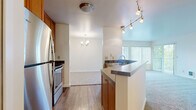 This single-floor residence offers a thoughtfully designed layout that balances functionality and comfort. The kitchen serves as a central hub, featuring warm wood cabinetry, stainless steel appliances, a dark laminate countertop peninsula with a built-in sink, and stylish track lighting overhead. The kitchen flows seamlessly into the dining area, which is accented by an elegant chandelier, and further opens to a bright living room bathed in natural light from a large sliding glass door. The living room provides direct access to a private balcony overlooking a lush, tree-lined outdoor setting. Soft carpeting runs throughout the living and bedroom areas, creating a warm and inviting atmosphere. The bedrooms are generously proportioned, each featuring large windows with vertical blinds that allow abundant natural light while maintaining privacy, along with baseboard heating for year-round comfort. The bathroom is finished with warm wood cabinetry, a stone-topped vanity, a large mirror, and durable hardwood-style flooring. Clean white walls and neutral tones throughout the residence create a bright, cohesive aesthetic that is both welcoming and easy to personalize.