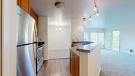 This single-floor residence offers a thoughtfully designed layout that balances functionality and comfort. The kitchen serves as a central hub, featuring warm wood cabinetry, stainless steel appliances, a dark laminate countertop peninsula with a built-in sink, and stylish track lighting overhead. The kitchen flows seamlessly into the dining area, which is accented by an elegant chandelier, and further opens to a bright living room bathed in natural light from a large sliding glass door. The living room provides direct access to a private balcony overlooking a lush, tree-lined outdoor setting. Soft carpeting runs throughout the living and bedroom areas, creating a warm and inviting atmosphere. The bedrooms are generously proportioned, each featuring large windows with vertical blinds that allow abundant natural light while maintaining privacy, along with baseboard heating for year-round comfort. The bathroom is finished with warm wood cabinetry, a stone-topped vanity, a large mirror, and durable hardwood-style flooring. Clean white walls and neutral tones throughout the residence create a bright, cohesive aesthetic that is both welcoming and easy to personalize.