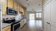 This single-floor residence offers a thoughtfully designed open-concept layout that seamlessly connects the kitchen and living area, creating a spacious and functional living environment. The kitchen is well-equipped with a full suite of stainless steel appliances, including a refrigerator, electric range, over-the-range microwave, and dishwasher. Light wood cabinetry pairs with granite-style countertops and a classic white tile backsplash, while track lighting overhead adds both practicality and a modern touch. Rich, dark hardwood-style flooring runs throughout the main living areas, lending warmth and continuity to the space. The living area is bright and open, benefiting from an abundance of natural light streaming through large windows and a sliding glass door that leads to a private balcony. A ceiling fan adds comfort to the living space. The bedroom area features built-in shelving and ample closet space. The bathroom is finished with matching cabinetry and granite-style countertops, complemented by light tile flooring, a large mirror, and convenient access to a walk-in closet. Neutral wall tones throughout create a clean, versatile backdrop suited to a variety of interior styles.