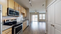 This single-floor residence offers a thoughtfully designed open-concept layout that seamlessly connects the kitchen and living area, creating a spacious and functional living environment. The kitchen is well-equipped with a full suite of stainless steel appliances, including a refrigerator, electric range, over-the-range microwave, and dishwasher. Light wood cabinetry pairs with granite-style countertops and a classic white tile backsplash, while track lighting overhead adds both practicality and a modern touch. Rich, dark hardwood-style flooring runs throughout the main living areas, lending warmth and continuity to the space. The living area is bright and open, benefiting from an abundance of natural light streaming through large windows and a sliding glass door that leads to a private balcony. A ceiling fan adds comfort to the living space. The bedroom area features built-in shelving and ample closet space. The bathroom is finished with matching cabinetry and granite-style countertops, complemented by light tile flooring, a large mirror, and convenient access to a walk-in closet. Neutral wall tones throughout create a clean, versatile backdrop suited to a variety of interior styles.