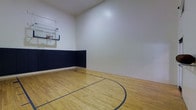 This impressive indoor basketball court offers a purpose-built athletic space designed for serious play and recreational enjoyment alike. The court features a classic hardwood floor finished in a warm natural tone, complete with regulation court lines and a three-point arc painted in a contrasting dark color for clear on-court orientation. A wall-mounted, adjustable glass backboard with a regulation hoop anchors one end of the court, providing an authentic game-ready setup. The lower portion of the surrounding walls is lined with navy blue padded wall padding, a practical safety feature that protects players during active play. Above the padding, the walls transition to a clean white finish that extends up to the high ceiling, contributing to the open and airy feel of the space. The generous ceiling height accommodates full-range shooting and movement without restriction. The overall design is clean, functional, and well-maintained, making this space equally suited for casual pickup games, personal training sessions, or skill development. The single-floor layout ensures straightforward accessibility throughout the court area.
