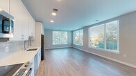 Located at 2008–2032 N Beauregard St in Alexandria, Virginia, this modern single-floor residence offers a thoughtfully designed layout with clean lines and contemporary finishes throughout. The open-concept living and dining area is bathed in natural light from a generous bank of large windows fitted with blinds, overlooking lush tree views. Light-toned hardwood-style flooring runs seamlessly through the main living spaces, creating a cohesive and airy feel. The kitchen is well-appointed with crisp white cabinetry extending to the ceiling, a classic subway tile backsplash, white quartz countertops, a stainless steel sink, and a full suite of stainless steel appliances including a refrigerator, range, dishwasher, and built-in microwave. A standout architectural detail is the set of frosted-glass sliding barn doors, which add a stylish, modern touch while defining the bedroom areas. The bedrooms are spacious and serene, with neutral tones and access to closet space. Two bathrooms serve the unit: one features a sleek, elongated vanity with a large mirror and a glass-enclosed walk-in shower with tile surround, while the second includes a full tub-shower combination with tiled walls. Recessed lighting throughout enhances the bright, contemporary ambiance of this well-finished Alexandria residence.