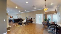 This single-story clubhouse offers a spacious and inviting communal environment designed for relaxation and social gathering. The main living area features warm hardwood-style flooring that runs throughout, creating a cohesive and polished aesthetic. Crown molding lines the ceilings, adding a refined architectural detail that enhances the overall elegance of the space.

The layout is open and generously proportioned, accommodating multiple seating arrangements that include plush sofas, accent chairs, and ottoman footrests arranged atop a decorative area rug. Recessed lighting and ceiling fans are evenly distributed across the ceiling, ensuring both comfort and ample illumination throughout the room.

A decorative buffet cabinet serves as a functional focal point, complemented by a coffee station nearby — a thoughtful amenity for residents and guests. A pendant light fixture adds a warm, inviting glow to the dining area, where a wooden table and chairs provide an additional gathering space.

Natural light filters into the room through a set of French doors featuring an arched transom window, connecting the interior to the outdoors. Neutral taupe walls and tasteful wall décor contribute to the warm, welcoming ambiance. Overall, this clubhouse presents a well-appointed and functional space suited for community events and everyday socializing.