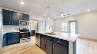 Located at 2422 S Carrier Pkwy in Grand Prairie, TX, this well-appointed 2-bedroom, 2-bathroom residence offers a thoughtfully designed single-level layout. The open-concept kitchen and living area serves as the heart of the home, anchored by a large central island with a white quartz countertop, undermount sink, and pendant lighting. Navy blue shaker-style cabinetry lines the kitchen walls, complemented by a subway tile backsplash, stainless steel appliances — including a refrigerator, range, microwave, and dishwasher — and light wood-tone flooring that extends throughout the main living areas. A ceiling fan overhead and windows offering outdoor views complete the living space. An in-unit washer and dryer are conveniently tucked within the kitchen area. Both bedrooms feature plush carpeting, ceiling fans, and generous windows that bring in natural light, along with dedicated closets for ample storage. The two bathrooms continue the navy cabinetry theme, featuring wood-tone flooring, white quartz vanity surfaces, brushed nickel fixtures, and glass-enclosed showers. The unit's clean, contemporary aesthetic is carried consistently throughout, creating a cohesive and modern living environment.