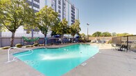 Located in Memphis, Tennessee, this outdoor pool area offers a well-appointed recreational space set against the backdrop of a multi-story building. The generously sized swimming pool features clear turquoise water with lane dividers and reaches a depth of 9 feet, accommodating both casual swimmers and more experienced guests. The pool's geometric layout provides ample swimming room, with wide concrete pool decking surrounding the perimeter for easy access via built-in entry steps and handrails.

The poolside area is designed for relaxation and social gathering, furnished with multiple seating arrangements including umbrella-shaded tables and chairs, providing comfortable outdoor lounging under open skies. Mature trees line portions of the enclosure, offering natural shade and a pleasant, leafy ambiance that softens the urban surroundings.

The entire pool area is enclosed by a secure iron perimeter fence, ensuring privacy and safety. Safety equipment, including a life ring station, is visibly mounted within the enclosure. The space benefits from abundant natural light throughout the day, with clear sightlines to the adjacent high-rise building and surrounding greenery. Overall, the pool area presents a functional and inviting outdoor amenity well-suited for both leisure and recreation in the heart of Memphis.