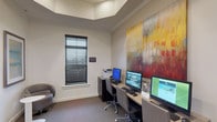 This single-floor business center is thoughtfully designed to balance functionality with a polished aesthetic. The room is anchored by a row of three desktop computer workstations arranged along a continuous desk surface, each equipped with an ergonomic rolling chair, keyboard, and mouse, making the space well-suited for productivity. A multifunction printer is conveniently positioned at the end of the workstation area.

A striking large-scale abstract painting in bold reds, yellows, and whites commands attention on the wall above the workstations, adding a vibrant artistic focal point to the otherwise neutral palette of soft gray walls and carpeted flooring. The elevated tray ceiling enhances the sense of vertical space and contributes to the room's refined character.

Natural light filters in through windows fitted with dark horizontal blinds, offering adjustable light control. A set of elegant white French doors with multi-pane glass panels provides both a visual connection to the adjacent space and an openness to the room. The lounge area features modern rounded armchairs in gray upholstery paired with sleek white pedestal side tables, offering a comfortable spot for informal conversations. Framed artwork and a tall decorative plant complete the welcoming, professional atmosphere.