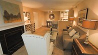 This single-floor space is designed with an open-concept layout that seamlessly connects the living area, dining nook, and kitchen, creating a cohesive and functional flow. The living area is generously sized, accommodating a seating arrangement that includes a studded white accent chair, a gray sofa, and a recliner, all centered around a light-toned coffee table. A decorative fireplace anchors one side of the room, complemented by a stylish wine rack that adds character. Warm, neutral-toned walls and carpeted flooring contribute to the inviting ambiance throughout. A sunburst mirror serves as a focal decorative element on the main wall, adding visual interest. Toward the back, a raised dining area features bar-height seating, offering a casual dining option. The kitchen is partially open to the main living space, featuring light cabinetry and overhead task lighting, making it feel integrated yet distinct. Track lighting and strategically placed lamps provide layered illumination throughout the space, enhancing the warm, cozy atmosphere. Overall, the space presents a comfortable and thoughtfully arranged interior with a consistent, welcoming aesthetic.