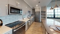 Located at 2 Carlson Ct in Charleston's historic 29403 zip code, this single-floor unit blends industrial character with contemporary design. The open-concept kitchen is a standout, outfitted with high-gloss grey cabinetry, white subway tile backsplash, light quartz countertops, and a full suite of stainless steel appliances. A large central island topped with a grey quartz surface is illuminated by a trio of brushed metal pendant lights, making it both a functional workspace and a natural gathering point. Light wood-look flooring flows throughout the main living areas, complementing the clean, modern palette. The living area features a striking exposed concrete ceiling with a ceiling fan, paired with a bold grid-pane window that draws in natural light and frames views of the surrounding neighborhood. The bathroom is finished with large-format marble-look wall tiles in the glass-enclosed shower, dark porcelain floor tiles, and crisp white walls, creating a clean and sophisticated feel. Grey paneled interior doors are used consistently throughout, reinforcing the cohesive, modern aesthetic. The overall ambiance is urban and refined, with industrial-inspired details softened by contemporary finishes throughout.