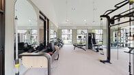 This fitness center is designed to provide a premium workout experience within a generously proportioned, single-floor layout. The space is divided into distinct functional zones, each thoughtfully outfitted to support a wide variety of training styles.

Architecturally, the facility features soaring vaulted ceilings — partially clad in warm wood paneling in one section — which lend visual interest and an open, airy quality throughout. Large floor-to-ceiling windows line multiple walls, flooding the interior with natural light and offering views of the surrounding exterior. Full-length mirrors further amplify the sense of space.

The cardio zone includes treadmills, stair climbers, ellipticals, rowing machines, and connected spin bikes equipped with individual screens. A large mounted display provides additional entertainment options. The strength training area features a multi-station functional rig with a punching bag, gymnastic rings, and cable attachments, alongside weight benches, selectorized machines, and a full dumbbell rack. Pilates-style reformer equipment is also present, supporting low-impact and rehabilitative training.

Flooring transitions between cushioned carpet and rubber matting in high-impact areas. The overall ambiance is clean, modern, and well-lit, creating an inviting environment for focused training.