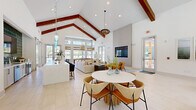 This single-floor community space in Savannah, Georgia offers a generous, open-concept layout defined by striking architectural character and a cohesive coastal-modern design. The most commanding feature is the dramatic vaulted ceiling adorned with rich, exposed wood beams that draw the eye upward and lend warmth to the otherwise crisp, white interior. Light-toned large-format tile flooring runs throughout, enhancing the sense of openness and continuity across the space.

Natural light floods the interior through an abundance of windows and glass doors positioned along multiple walls, offering views of the surrounding outdoor greenery. The main gathering area features a long bar-height counter with navy upholstered stools, ideal for casual seating. Pendant lighting in aqua glass and woven rattan styles adds a distinctive coastal flair overhead.

A lounge area is anchored by plush neutral sectional sofas and accented by mustard-toned armchairs positioned near a wall-mounted flat-screen display. A separate seating vignette with a round white table and woven chairs provides additional gathering space. A sleek glass-paneled barn door separates zones within the layout while maintaining visual openness. Coastal-themed artwork, lush greenery, and layered textures throughout complete the inviting, resort-inspired ambiance.