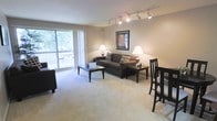 This single-floor space offers a well-organized, open-concept layout that seamlessly integrates the living and dining areas into one cohesive environment. The living area is carpeted throughout and anchored by a comfortable sofa, a rectangular coffee table, and tasteful wall art, all bathed in warm light from twin table lamps and a modern track lighting system mounted on the ceiling. The track lighting provides flexible, directional illumination across the room, complementing the natural light that streams in through a large sliding glass door. The dining area is positioned adjacent to the living space, featuring a dark-toned table and matching chairs that coordinate well with the overall color scheme of rich browns and neutral beige tones. The large window and sliding glass door draw the eye outward to a balcony, offering a glimpse of lush greenery beyond—adding a refreshing natural element to the interior ambiance. The neutral wall tones and light carpet create a clean, inviting atmosphere throughout. The overall design is practical and comfortable, making efficient use of the available space while maintaining an open, airy feel suitable for both relaxing and entertaining.