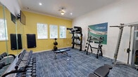 Located at 12055 Sabo Rd in Houston, TX, this single-room fitness center offers a well-equipped workout environment designed to support a variety of exercise routines. The space features crown molding along the ceiling and is finished with a durable, patterned blue carpet flooring suited for high-traffic use. The room is divided into distinct functional zones: one side is painted a warm yellow and lined with windows fitted with white blinds, allowing natural light to fill the space and offering views of surrounding greenery. This area houses a weight plate storage rack and TRX suspension training equipment. The opposite side, finished in crisp white, features a large wall-mounted mirror that enhances the sense of space, alongside a dumbbell rack stocked with a full range of free weights. A cable machine and additional free weight storage racks provide strength training versatility. Multiple flat-screen TVs are mounted on the walls, keeping users entertained during workouts. Adjustable workout benches are positioned throughout the floor, accommodating both pressing and incline exercises. Recessed lighting and a ceiling fan ensure the room stays well-lit and comfortable. A small towel and accessory station near the entrance adds a convenient amenity touch. The overall ambiance is functional, clean, and purposefully outfitted for a complete strength and conditioning experience.