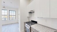 Located at 118–126 S 10th St in Newark, NJ, this efficiently designed unit offers a thoughtfully arranged single-floor layout comprising a kitchen and a full bathroom. Light wood-style luxury vinyl flooring runs throughout the main living area and kitchen, lending a clean, cohesive aesthetic to the space.

The kitchen is well-appointed with stainless steel appliances, including a gas range with a range hood, a refrigerator, and a double-basin sink. Wood-toned lower cabinetry pairs with crisp white upper cabinets and a classic subway tile backsplash, while granite countertops provide a polished, durable work surface. Recessed lighting illuminates the kitchen efficiently.

The main living area benefits from two large, grid-pane windows fitted with black frames that flood the room with natural light and offer views of the surrounding Newark neighborhood. A pendant light fixture adds a stylish focal point to the bright, open space, complemented by clean white walls and tall baseboards.

The bathroom features a refined tile design, with large-format light gray wall tiles accented by wood-toned vertical tile detailing. The floor incorporates a striking mix of gray and wood-look tiles in a horizontal pattern. Black matte hardware and fixtures add a modern contrast, completing a bathroom that is both functional and visually appealing.