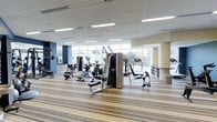 This fitness studio is a thoughtfully designed, single-floor facility that offers a comprehensive exercise environment for users of all fitness levels. The space is characterized by its bright, open layout, with floor-to-ceiling windows along one wall that flood the interior with abundant natural light and provide views of the surrounding greenery outdoors.

The studio is outfitted with a diverse selection of strength training equipment, including multi-station cable machines, adjustable weight benches, and free weights, catering to a full range of resistance training needs. Cardio equipment, such as treadmills and stationary bikes, is positioned near the windows to take advantage of the natural light and outdoor views, creating an energizing atmosphere for endurance workouts.

The flooring consists of a striped, cushioned carpet that adds comfort underfoot while providing a clean, modern aesthetic. Overhead, recessed linear LED lighting supplements the natural light, ensuring the space remains well-lit throughout. The neutral color palette of beige and white walls, accented by dark navy blue panels, contributes to a professional and focused ambiance. The open floor plan allows for easy navigation between equipment zones, making this fitness studio a functional and inviting space for daily exercise.