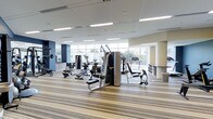 This fitness studio is a thoughtfully designed, single-floor facility that offers a comprehensive exercise environment for users of all fitness levels. The space is characterized by its bright, open layout, with floor-to-ceiling windows along one wall that flood the interior with abundant natural light and provide views of the surrounding greenery outdoors.

The studio is outfitted with a diverse selection of strength training equipment, including multi-station cable machines, adjustable weight benches, and free weights, catering to a full range of resistance training needs. Cardio equipment, such as treadmills and stationary bikes, is positioned near the windows to take advantage of the natural light and outdoor views, creating an energizing atmosphere for endurance workouts.

The flooring consists of a striped, cushioned carpet that adds comfort underfoot while providing a clean, modern aesthetic. Overhead, recessed linear LED lighting supplements the natural light, ensuring the space remains well-lit throughout. The neutral color palette of beige and white walls, accented by dark navy blue panels, contributes to a professional and focused ambiance. The open floor plan allows for easy navigation between equipment zones, making this fitness studio a functional and inviting space for daily exercise.