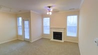 This single-floor space centers around a welcoming living area designed for comfort and functionality. The room features a classic fireplace with a white-painted mantel, serving as the focal point of the space and adding a warm, inviting ambiance. Crown molding runs along the perimeter of the ceiling, lending a refined architectural detail to the otherwise understated design. A ceiling fan with an integrated light fixture is centrally mounted, ensuring year-round climate comfort and ambient lighting. The room is finished with wall-to-wall carpeting in a neutral beige tone, contributing to a soft, comfortable feel underfoot. Natural light is generously supplied through multiple windows fitted with horizontal blinds, allowing for adjustable light control throughout the day. A glass-paneled exterior door on one side provides additional natural light and potential outdoor access. The walls are painted in a warm, neutral tone that complements the carpeting and white trim, creating a cohesive and airy aesthetic. Overall, the space presents a clean, move-in-ready environment with classic detailing and practical features well-suited for everyday living.