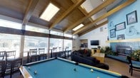 This inviting clubroom is thoughtfully designed to serve as a versatile social hub. The space is immediately distinguished by its striking vaulted wood-beam ceiling, which adds warmth and architectural character while creating a sense of openness and volume. Flush skylight panels integrated into the ceiling provide consistent, even illumination throughout the day, complementing the generous natural light that floods in through a full wall of large windows along one side of the room.

At the heart of the space sits a full-sized billiards table, making it an ideal spot for recreation and casual entertainment. Adjacent to it, a comfortable lounge area features a plush sectional sofa and accent furnishings arranged around a flat-screen television, perfect for unwinding or watching a game. High-top bar seating positioned along the window wall offers additional gathering space with pleasant outdoor views of the surrounding greenery.

A built-in kitchenette is conveniently tucked into the background, equipped with cabinetry and appliances to support light refreshments and entertaining needs. Lush indoor plants are scattered throughout, adding a refreshing, natural touch to the overall ambiance. The combination of warm wood tones, bright open windows, and thoughtfully arranged amenities makes this clubroom a welcoming and functional retreat.