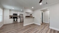 This single-floor space showcases a clean, contemporary design with consistent warm-toned luxury vinyl plank flooring and crisp white trim running throughout. The open-concept layout connects the kitchen and living area seamlessly, creating a bright and airy atmosphere enhanced by recessed lighting and ceiling fans.

The kitchen is a standout feature, outfitted with white shaker-style cabinetry extending to the ceiling for ample storage, warm wood-toned countertops, a neutral tile backsplash, and a full suite of stainless steel appliances including a refrigerator, gas range, microwave, and dishwasher. A peninsula with a built-in upper cabinet and a lower breakfast bar creates a natural divider between the kitchen and the open living space, offering additional functionality without sacrificing the open feel.

The bathroom is thoughtfully designed with a white vanity cabinet topped with a rich brown countertop, a framed mirror, and a glass-enclosed walk-in shower with white tile surround. A window with plantation shutters brings in natural light while maintaining privacy.

The bedroom is a well-proportioned room featuring a double-door closet with dual hanging rods, offering generous storage capacity. Crown molding details and large baseboards add a refined architectural touch throughout the space, lending a polished, move-in-ready character to this well-designed unit.